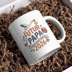 Peut inclure: Mug en céramique blanche avec l'inscription "FUTUR PAPA 2026" en orange et bleu, représentant une cigogne portant un bébé. La tasse comprend également une pipe, une boîte à outils et des chaussons.