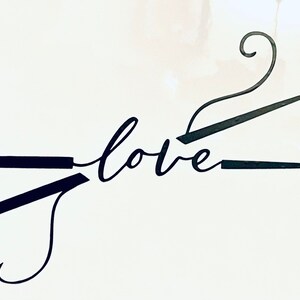 May include: Black metal art piece featuring knitting needles and a crochet hook forming the word "love" in cursive script. The needles and hook are set against a white background, creating a minimalist design.