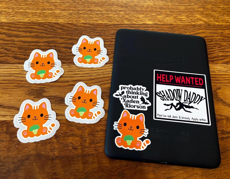 May include: A collection of orange cat stickers with green accents and a black e-reader with stickers. The e-reader has stickers that read "Help Wanted Shadow Daddy" and "probably thinking about Xaden Riorson."