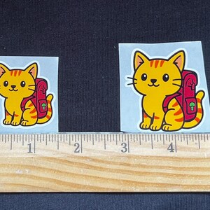 Puede incluir: Dos pegatinas con un gato de dibujos animados con una mochila. El gato es amarillo con rayas naranjas y una mochila roja. Una regla de madera se coloca debajo.