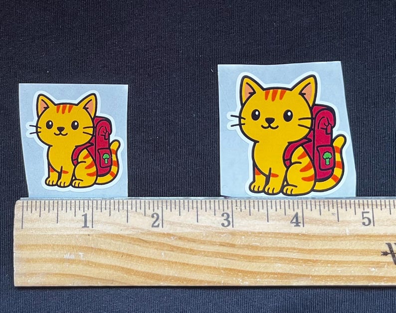 Puede incluir: Dos pegatinas con un gato de dibujos animados con una mochila. El gato es amarillo con rayas naranjas y una mochila roja. Una regla de madera se coloca debajo.