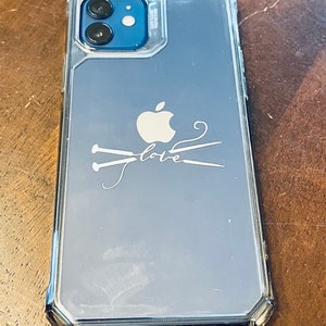 May include: Clear phone case with a silver border, featuring an Apple logo and the word "love" with knitting needles. The case is designed to fit a smartphone with a dual-lens camera.