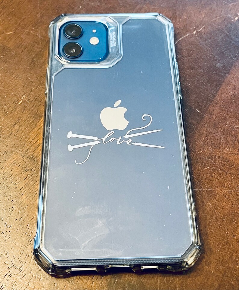 May include: Clear phone case with a silver border, featuring an Apple logo and the word "love" with knitting needles. The case is designed to fit a smartphone with a dual-lens camera.
