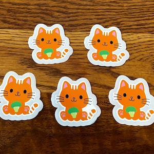 May include: Five orange cat stickers with black whiskers, a green object, and a white border are arranged on a wooden surface. The cartoon cats have a smiling face and are holding a green object.
