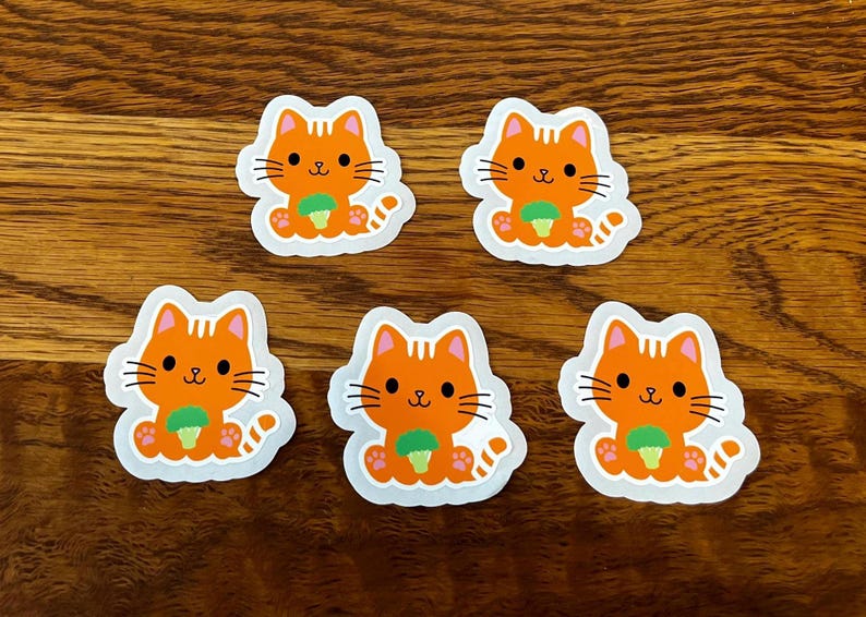 May include: Five orange cat stickers with black whiskers, a green object, and a white border are arranged on a wooden surface. The cartoon cats have a smiling face and are holding a green object.