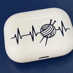 May include: White AirPod case with a black graphic of a yarn ball and knitting needles integrated into a heartbeat line. The case is on a dark blue surface.
