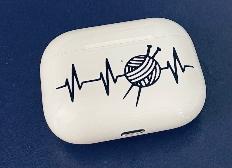 May include: White AirPod case with a black graphic of a yarn ball and knitting needles integrated into a heartbeat line. The case is on a dark blue surface.