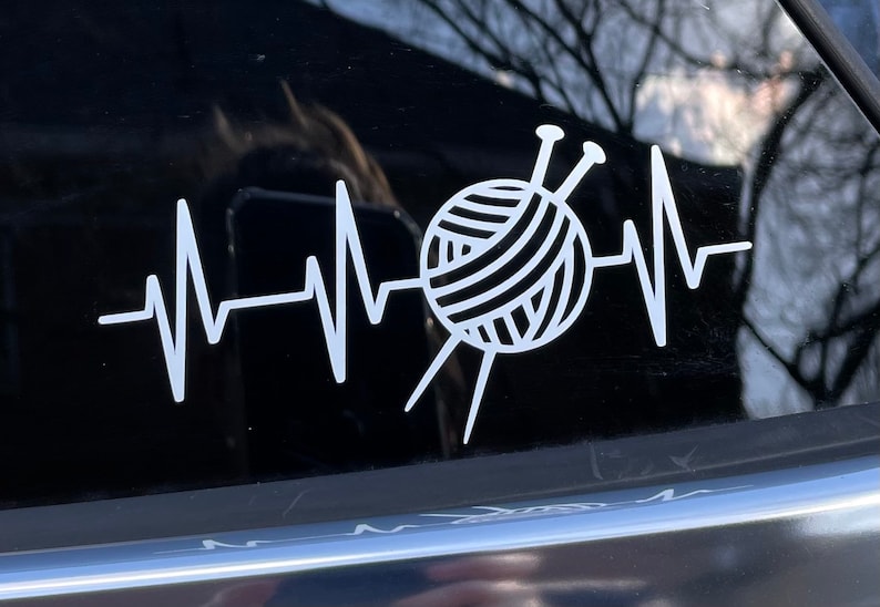 May include: White vinyl decal featuring a heartbeat design with a yarn ball and knitting needles in the center. The design is a visual representation of the love for knitting and crafting. The decal is applied to a dark surface.