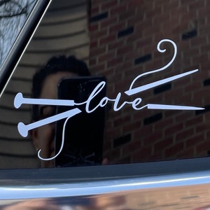 May include: White vinyl decal with the word "love" and a knitting needle design. The decal is applied to a car window, with reflections visible. The design is simple and elegant, perfect for knitters.