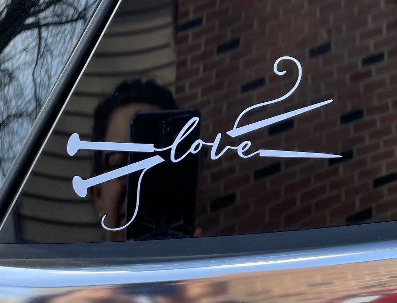May include: White vinyl decal with the word "love" and a knitting needle design. The decal is applied to a car window, with reflections visible. The design is simple and elegant, perfect for knitters.