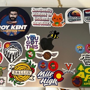 May include: A laptop covered in colorful stickers. Stickers include a cartoon cat, a bee over an apple, and text that reads "ROY KENT" and "YOU GOT THIS". Other stickers feature Colorado and "Mile High" designs.