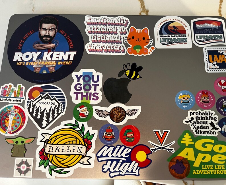 May include: A laptop covered in colorful stickers. Stickers include a cartoon cat, a bee over an apple, and text that reads "ROY KENT" and "YOU GOT THIS". Other stickers feature Colorado and "Mile High" designs.