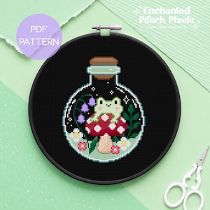 May include: A cross-stitch pattern in a black embroidery hoop featuring a light green frog sitting on a red and white spotted mushroom inside a glass bottle. The design includes purple lily of the valley flowers, small white flowers, and green leaves.