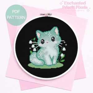 Könnte beinhalten: Ein Kreuzstichmuster mit einer hellblauen Katze mit weißen Akzenten, die auf grünem Gras vor schwarzem Hintergrund sitzt. Das Design ist in einem weißen Stickrahmen gerahmt, mit dem Text "PDF PATTERN" und "Enchanted Peach Pixels".