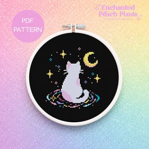 May include: A white embroidery hoop containing a cross stitch pattern on black fabric. The design features a white cat sitting on a colourful swirling base, surrounded by yellow stars and a yellow crescent moon. Text reads PDF PATTERN.