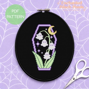 Peut inclure: Un projet de broderie au point de croix dans un tambour circulaire noir, représentant un cercueil aux contours violets contenant des fleurs de muguet en forme de fantômes blancs, des feuilles vertes et un croissant de lune jaune sur fond noir.