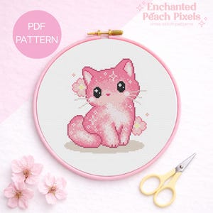 May include: A pink cross-stitch cat design in a pink embroidery hoop. The text "PDF PATTERN" is in a pink circle. The text "Enchanted Peach Pixels" is at the top. Yellow scissors and pink flowers are also in the image. The design is a PDF pattern.