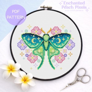 Puede incluir: Un patrón de punto de cruz de una polilla verde y turquesa con detalles de luna, rodeada de flores rosas y moradas, en un aro negro. El texto incluye "PDF PATTERN" y "Enchanted Peach Pixels cross stitch patterns".