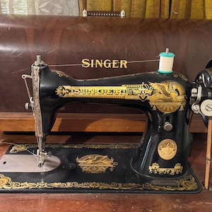 Könnte beinhalten: Vintage Singer Nähmaschine in Schwarz mit goldenen Akzenten. Die Maschine hat verzierte Designs, das Singer-Logo und eine Garnrolle. Sie steht auf einer Holzoberfläche, mit einem passenden Holzkoffer im Hintergrund.
