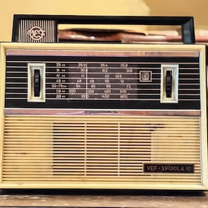 Könnte beinhalten: Vintage VEF Spīdola 10 Radio mit cremefarbener Front und schwarzem Oberteil. Das Radio hat ein Einstellrad mit Frequenzmarkierungen und zwei Bedienknöpfen. Der Text "VEF-SPIDOLA 10" ist sichtbar.