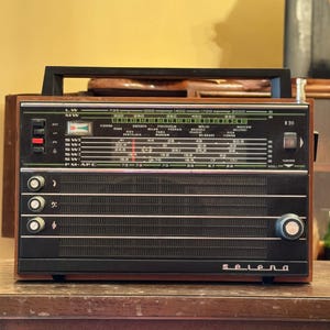 Op de afbeelding: Een vintage Selena-radio met een houten kast, zwarte voorkant en een handvat. De radio heeft een afstemknop, zenderindicatoren en de naam "selena" op de voorkant. Het heeft een retro-ontwerp.