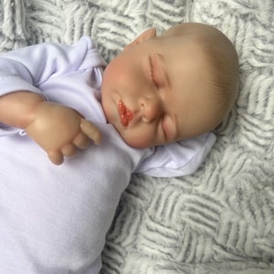 May include: A realistic baby doll with closed eyes and a slightly open mouth, dressed in a white onesie. The doll's skin has a lifelike tone, and it lies on a soft, textured, grey and white blanket. The doll's features are detailed.