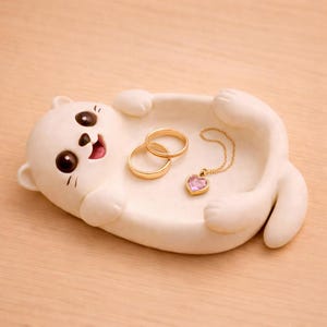 May include: A white ceramic cat-shaped trinket dish with two gold rings and a gold necklace with a pink heart-shaped pendant. The cat has large brown eyes and a smiling expression. The dish is on a light brown surface.