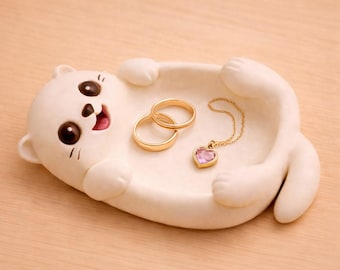 May include: A white ceramic cat-shaped trinket dish with two gold rings and a gold necklace with a pink heart-shaped pendant. The cat has large brown eyes and a smiling expression. The dish is on a light brown surface.
