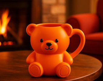 May include: An orange ceramic mug shaped like a teddy bear, with a black nose and eyes, and a handle. The mug is sitting on a dark wooden table. The background includes a fireplace and a red chair.