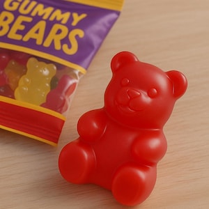 Könnte beinhalten: Ein leuchtend roter Gummibär-Bonbon liegt auf einer Holzoberfläche neben einer Tüte Gummibärchen. Die Tüte ist lila mit gelbem Text, der "Gummy Bears" lautet. Der Gummibär hat die klassische Bärenform.