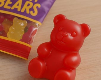 Puede incluir: Un caramelo de goma con forma de oso rojo brillante está sobre una superficie de madera junto a una bolsa de ositos de goma. La bolsa es morada con texto amarillo que dice "Gummy Bears". El osito de goma tiene la forma clásica de oso.