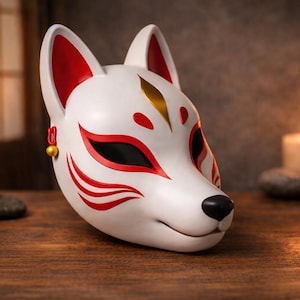 May include: A white fox mask with red and gold accents. The mask features red markings around the eyes and on the cheeks, with a black nose and ears lined with red. The mask is set on a wooden surface with candles in the background.
