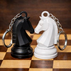 Peut inclure: Deux porte-clés en forme de cheval de jeu d'échecs, un noir et un blanc, sont présentés. Chaque porte-clés présente un design détaillé de tête de cheval et un porte-clés argenté. Les porte-clés sont posés sur un fond flou et un échiquier.