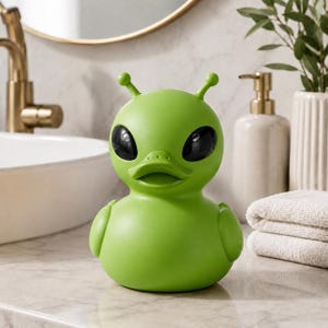 Puede incluir: Un pato de goma de color verde lima brillante con forma de alienígena, que presenta dos antenas en la parte superior de la cabeza, grandes ojos ovalados negros y lisos, y un pico de pato. La figura tiene un cuerpo redondeado con pequeñas alas.