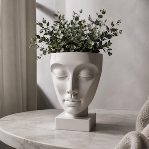 Pode incluir: Um vaso de cerâmica branco mate em forma de rosto humano com olhos fechados, nariz definido e lábios carnudos. O vaso repousa sobre uma base quadrada e está cheio de uma planta verde exuberante com folhagem pequena e arredondada.