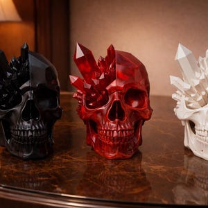Peut inclure: Trois sculptures décoratives en forme de crâne. L'une est noire avec des cristaux noirs, la seconde est rouge avec des cristaux rouges et la troisième est blanche avec des cristaux blancs. Les crânes sont exposés sur une surface sombre et réfléchissante.