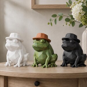 Può includere: Tre statuette geometriche di rane in bianco, verde e nero, ognuna con un cappello del colore corrispondente. Le rane presentano un design sfaccettato a basso poligono con superfici angolari distinte, disposte in fila su una superficie.