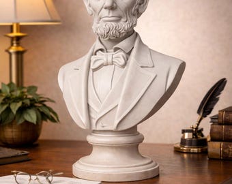 May include: A detailed bust of Abraham Lincoln, sculpted in a light, neutral tone. The sculpture features a bow tie and a formal suit. The bust is displayed on a wooden desk, alongside books, a lamp, and writing implements.