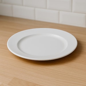 Puede incluir: Un plato redondo de cerámica blanca con un borde ligeramente elevado. Se encuentra sobre una superficie de madera clara, con azulejos blancos en el fondo. Este plato sencillo es adecuado para el uso diario y mide unos 25 cm de diámetro.
