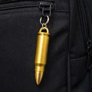May include: A gold-colored bullet-shaped keychain hanging from a black zipper. The keychain is made of metal and has a cylindrical body with a pointed tip. The background is a black fabric.