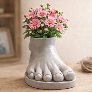 Puede incluir: Un jarrón gris de cerámica con forma de pie que contiene flores rosas y amarillas. El jarrón presenta un diseño detallado de un pie, incluyendo los dedos y una base redonda. Las flores están en plena floración, añadiendo un toque de color.
