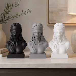 May include: Three sculptural busts of a person with long hair, each wearing a blindfold over their eyes. The figures are presented in a row, with one matte black, one matte gray, and one matte white bust, all mounted on square bases.