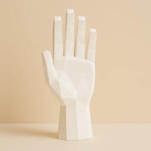Op de afbeelding: Een witte, geometrische handsculptuur met een facetontwerp. De sculptuur is een decoratief object, met een gestileerde open hand met uitgestrekte vingers. De basis heeft de vorm van een afgeknotte kegel. De achtergrond is effen en neutraal.