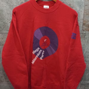 Op de afbeelding: Een felrood sweatshirt met ronde hals en een grote paarse cirkelvormige afbeelding op de borst. De afbeelding bevat de tekst "Virgin Atlantic" en "Mega Millennium Party" in witte en paarse letters. Een klein blauw logo staat op de linkermouw.