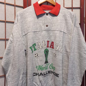 Puede incluir: Una sudadera gris de manga larga con cuello rojo. La parte delantera presenta el texto "ITALIA 90" en verde y rojo, junto con un trofeo verde y balones de f&uacute;tbol. Debajo est&aacute;n las palabras "World Cup CHALLENGE Football".