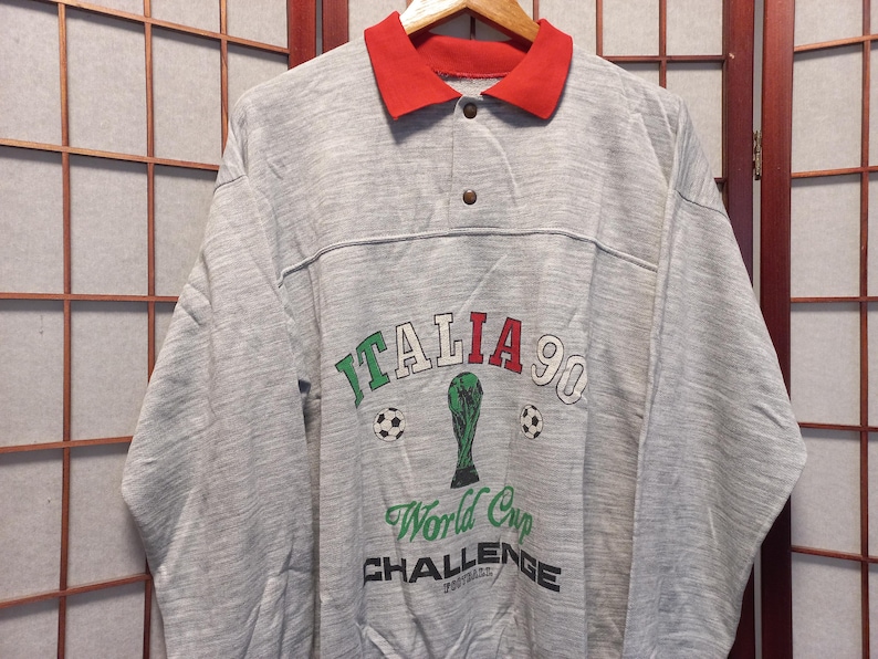 Puede incluir: Una sudadera gris de manga larga con cuello rojo. La parte delantera presenta el texto "ITALIA 90" en verde y rojo, junto con un trofeo verde y balones de f&uacute;tbol. Debajo est&aacute;n las palabras "World Cup CHALLENGE Football".