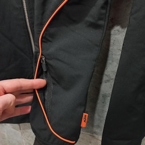 May include: Black jacket with orange trim and a zippered pocket. The jacket has a black ribbed cuff and a small orange KTM logo patch. The jacket is made of a smooth, possibly nylon, material.