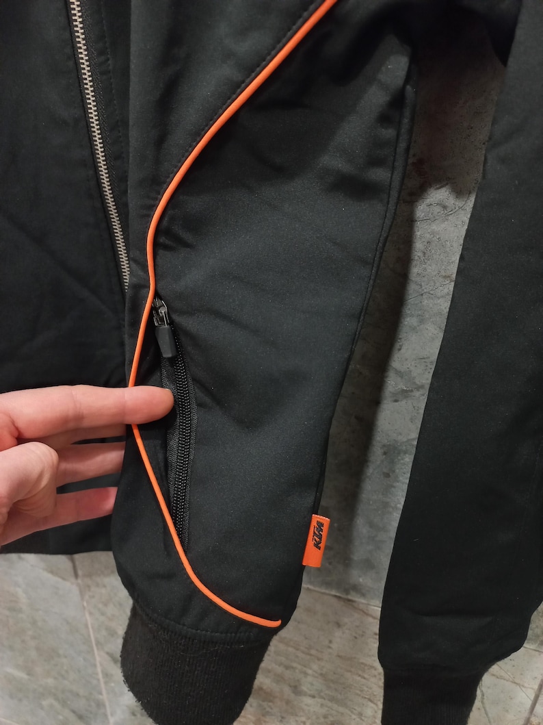 May include: Black jacket with orange trim and a zippered pocket. The jacket has a black ribbed cuff and a small orange KTM logo patch. The jacket is made of a smooth, possibly nylon, material.