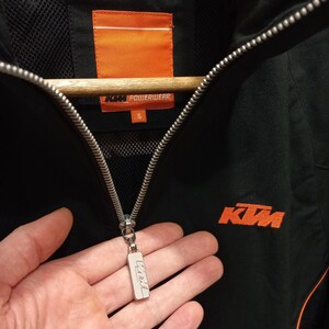 May include: Black KTM Powerwear jacket with orange accents. Features a full zipper closure and the KTM logo in orange on the chest. The jacket has a small size tag and a silver zipper pull.