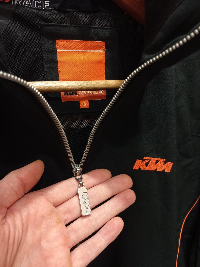 May include: Black KTM Powerwear jacket with orange accents. Features a full zipper closure and the KTM logo in orange on the chest. The jacket has a small size tag and a silver zipper pull.